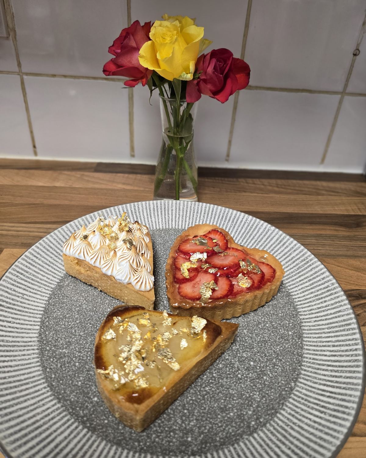 Three dessert tarts, lemon meringue, strawberry and gold leaf, with a small vase of roses